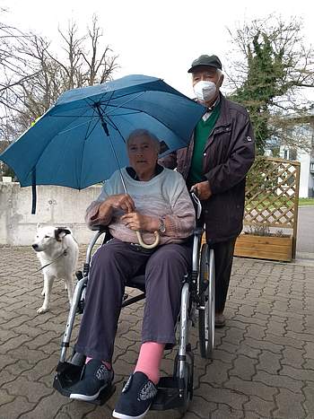 Ein Besucher schiebt eine Bewohnerin, die im Rollstuhl sitzt und daneben geht ein Hund.
