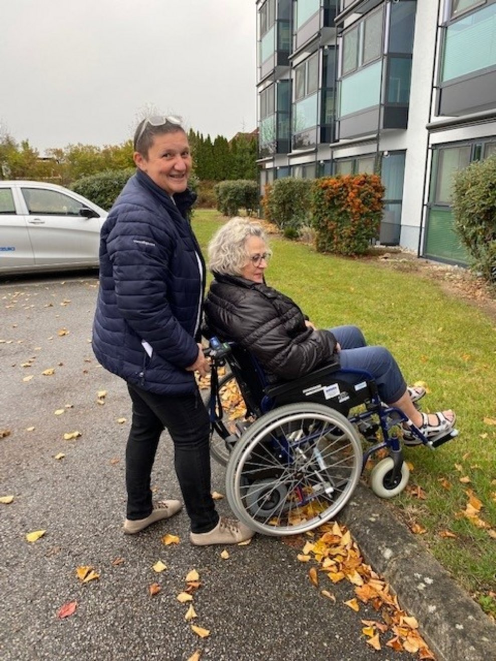 Mitarbeiterin schiebt andere Mitarbeiterin mit dem Rollstuhl